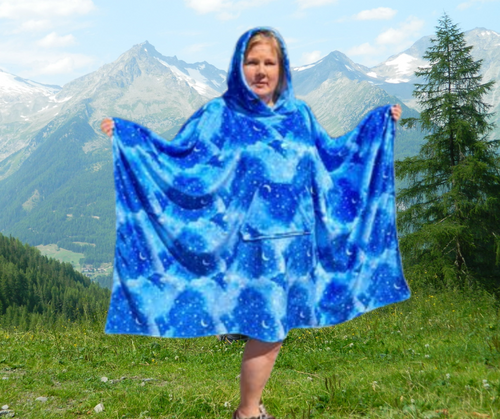 Woman wearing a hooded blanket made with a sky print fleece and front pocket with grass and mountain background